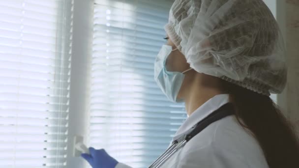 防護服を着た疲れた女医者が窓を開けて医者のマスクを外し疲れで目を閉じて新鮮な空気を吸う。病院での医師の勤務日。流行性コロナウイルス｜Covid19. — ストック動画