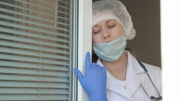 Dia de trabalho duro de médico no hospital. mulher cansada médico em terno protetor olha pela janela, tira sua máscara médica, fechando os olhos da fadiga, respira ar fresco. Coronovírus epidémico — Fotografia de Stock