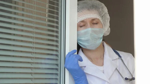 Dia de trabalho duro de médico no hospital. mulher cansada médico em terno protetor olha pela janela, tira sua máscara médica, fechando os olhos da fadiga, respira ar fresco. Coronovírus epidémico — Fotografia de Stock