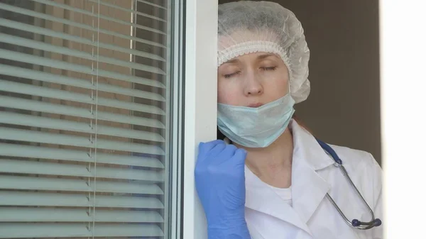 Hard working day of doctor in hospital. tired woman doctor in protective suit looks out window, takes off his medical mask, closing his eyes from fatigue, breathes fresh air. Epidemic Coronovirus