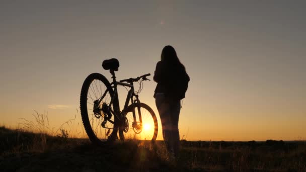 Retkeilijä terve nuori nainen seisoo mäellä vieressä polkupyörän, nauttia luonnosta ja auringosta. Vapaa tyttö matkustaa polkupyörän kanssa auringonlaskun aikaan. käsite seikkailu ja matka. yksinäinen nainen pyöräilijä lepää puistossa . — kuvapankkivideo