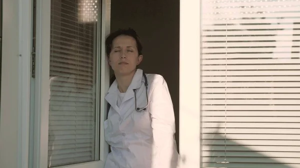 Zmęczona lekarka w kombinezonie ochronnym patrzy przez okno, zdejmuje maskę medyczną, zamyka oczy przed zmęczeniem, oddycha świeżym powietrzem. Ciężki dzień pracy lekarza w szpitalu. Pandemiczny koronawirus — Zdjęcie stockowe