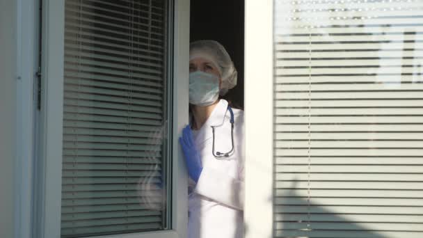 병원에서 힘든 하루를 보내고 있어요. 보호용 양복을 입은 피곤 한 여인의 의사는 창밖을 내다보고, 의료용 마스크를 벗고, 피로 에서 눈을 감고, 신선 한 공기를 호흡 한다. 유행하는 코로나 바이러스 — 비디오