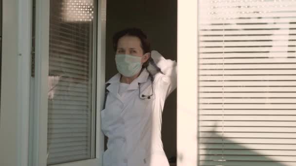 Μια κουρασμένη γιατρός με προστατευτική στολή κοιτάζει έξω από το παράθυρο, βγάζει την ιατρική του μάσκα, κλείνει τα μάτια του από την κούραση, αναπνέει καθαρό αέρα. Σκληρή μέρα εργασίας γιατρού στο νοσοκομείο. Πανδημία του Coronavirus — Αρχείο Βίντεο