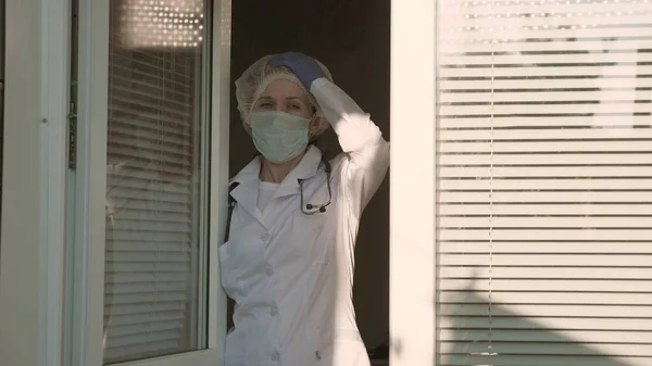 A tired woman doctor in protective suit looks out window, takes off his medical mask, closing his eyes from fatigue, breathes fresh air. Hard working day of doctor in hospital. Pandemic Coronavirus — Stock Photo, Image