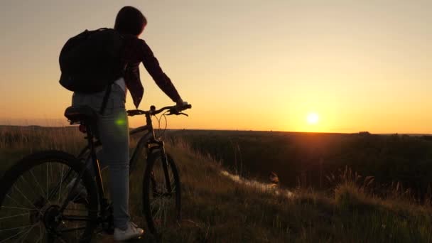 Caminhante mulher jovem saudável anda de bicicleta para a borda da colina, desfrutando da natureza e do sol. Menina livre viaja com uma bicicleta ao pôr do sol. conceito de aventura e viagens. mulher solitária ciclista descansando no parque . — Vídeo de Stock