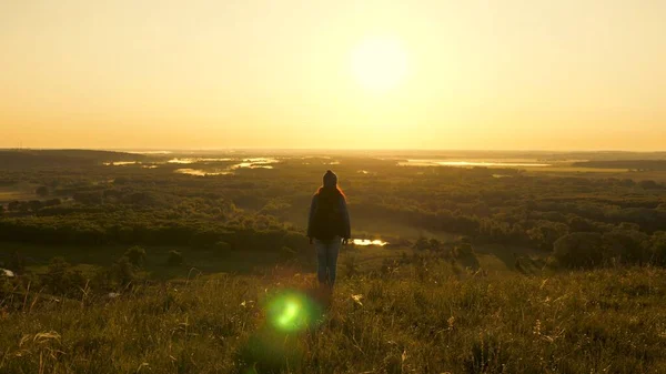 Una giovane viaggiatrice con uno zaino si trova ai margini della montagna nei raggi dell'alba, alza le mani in aria e gode della vittoria, del bel sole e del paesaggio. gratis turista ragazza viaggia da solo nel parco — Foto Stock