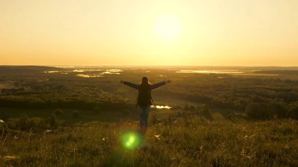 Una joven viajera con una mochila se encuentra al borde de la montaña en rayos del amanecer, levanta las manos en el aire y disfruta de la victoria, el hermoso sol y el paisaje. chica turística libre viaja solo en el parque —  Fotos de Stock