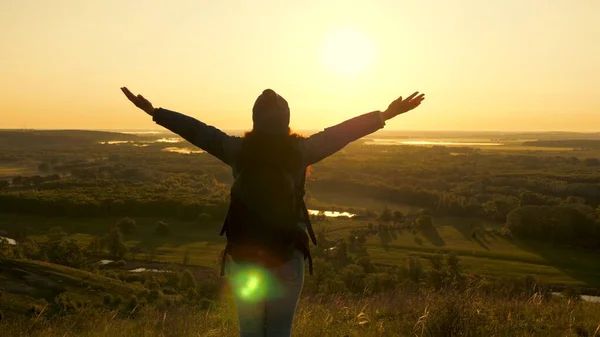 Una giovane viaggiatrice con uno zaino si trova ai margini della montagna nei raggi dell'alba, alza le mani in aria e gode della vittoria, del bel sole e del paesaggio. gratis turista ragazza viaggia da solo nel parco — Foto Stock