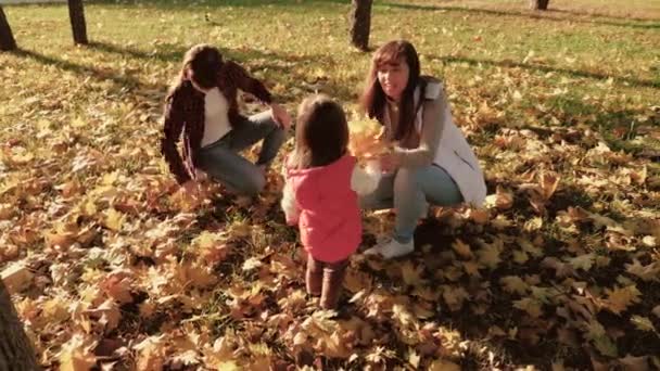 En ung mor leker med sina döttrar samlar fallna löv i en höstpark. familjen sitter på gula blad i trädgården. Begreppet moderskap i barndomen. lyckligt barn leker med mamma och syster. — Stockvideo