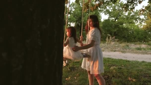 En mamma leker med barnet de svingar på rep på en ekgren i skogen. gratis familj har kul i Park. mor stenar frisk liten dotter på gunga under träd i solen. — Stockvideo