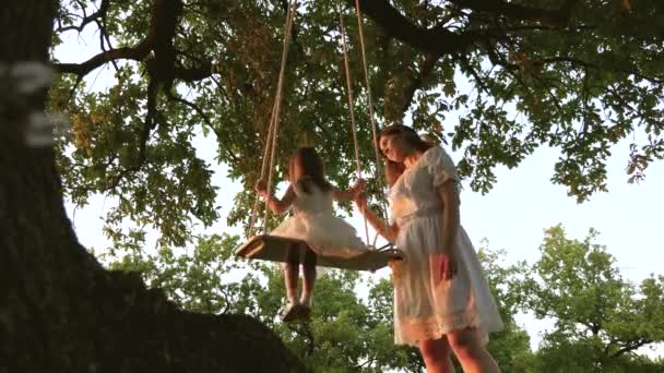 Mom shakes a healthy little daughter on swing under tree in sun. mother plays with child they are swinging on a rope on an oak branch in forest. girl laughs, rejoices. Free family having fun in park — Stock Video