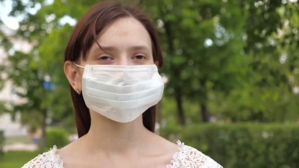 Maska ochronna na twarzy młodej dziewczyny w parku. Dziewczyna na ulicy miasta podróżuje w masce medycznej. turystka chroni twarz przed wirusami. Idź w masce na dół ulicy do sklepu — Wideo stockowe