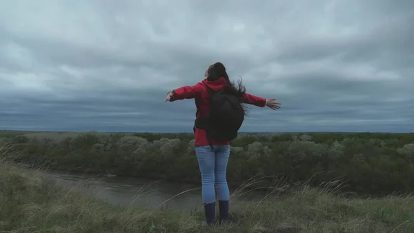 free happy girl traveler goes to edge of mountain with outstretched arms, wind is waving her hair. young woman travels with a backpack, looks from top of mountain, enjoying landscape and river