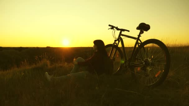 Unavená cyklistka, která večer odpočívá na okraji útesu a obdivuje slunce. Volná dívka cestuje s bicyklem při západu slunce. Zdravá mladá žena turistka sedí na kopci vedle kola, těší příroda a slunce — Stock video