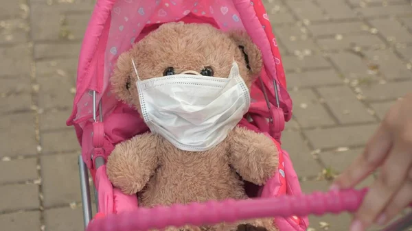 Niño en la calle con su juguete favorito en una máscara protectora. concepto de infancia saludable. niña camina en el parque con un cochecito y un osito de peluche con máscara médica. niño juega una epidemia y protege el juguete. — Foto de Stock