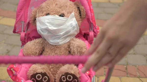 Niño en la calle con su juguete favorito en una máscara protectora. concepto de infancia saludable. niña camina en el parque con un cochecito y un osito de peluche con máscara médica. niño juega una epidemia y protege el juguete. — Foto de Stock