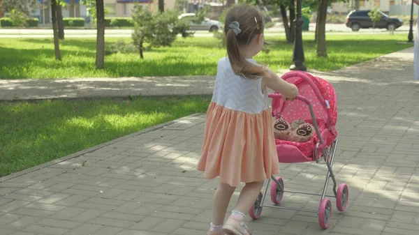 Niña camina en el parque con un cochecito y un osito de peluche con máscara médica. niño juega una epidemia y protege el juguete. niño en la calle con su juguete favorito en una máscara protectora. concepto de infancia saludable — Foto de Stock