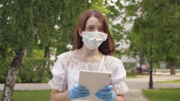 Ein gesundes Mädchen läuft mit Schutzmaske und Handschuhen im Stadtpark und arbeitet mit Tabletten. COVID-19. Virengefahr. junge Geschäftsfrau, die auf der Straße arbeitet. Sichere und soziale Distanz. — Stockvideo