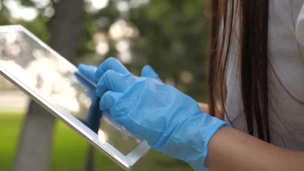 Een gezond meisje loopt in medische handschoenen met een tablet in een stadspark. close-up. COVID-19. gevaar voor het virus. jonge zakenvrouw die op afstand op straat werkt. Veilige en sociale afstand. — Stockvideo