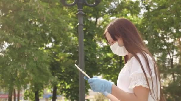 Ein gesundes Mädchen läuft mit Schutzmaske und Handschuhen im Stadtpark und arbeitet mit Tabletten. COVID-19. Virengefahr. junge Geschäftsfrau, die auf der Straße arbeitet. Sichere und soziale Distanz. — Stockvideo