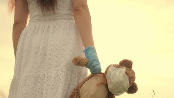 Девушка носит в руке игрушечного плюшевого мишку в медицинской маске в летнем парке. ребенок держит своего любимого плюшевого мишку в защитной маске во время карантина. Пандемический коронавирус Ковид-19. концепция детского здоровья — стоковое видео