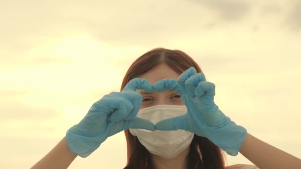 Fille dans un masque de protection et des gants médicaux montre le symbole du cœur avec ses mains. jeune femme aime les médecins. pandémie, covide 19. femme médecin montrant les doigts du cœur et l'amour pour les patients. — Video