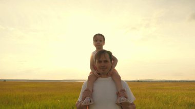 Baba gün batımında kızıyla birlikte omuzlarında yürüyor. Babam sevgili çocuğunun omuzlarını güneş ışınlarıyla taşır. Ailesi olan çocuk gün batımında yürür. Mutlu aile parkta dinleniyor..