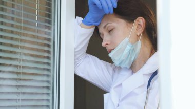Koruyucu takım elbiseli yorgun bir kadın doktor pencereye yaklaşıyor, tıbbi maskesini ve şapkasını çıkarıyor, gözlerini yorgunluktan kapıyor, temiz hava soluyor. Hastanede çok çalışan bir doktor var..