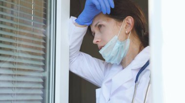 Koruyucu takım elbiseli yorgun bir kadın doktor pencereye yaklaşıyor, tıbbi maskesini ve şapkasını çıkarıyor, gözlerini yorgunluktan kapıyor, temiz hava soluyor. Hastanede çok çalışan bir doktor var..
