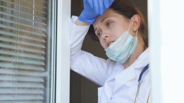 Koruyucu takım elbiseli yorgun bir kadın doktor pencereye yaklaşıyor, tıbbi maskesini ve şapkasını çıkarıyor, gözlerini yorgunluktan kapıyor, temiz hava soluyor. Hastanede çok çalışan bir doktor var..