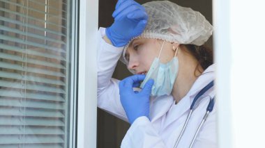 Koruyucu takım elbiseli yorgun bir kadın doktor pencereye yaklaşıyor, tıbbi maskesini ve şapkasını çıkarıyor, gözlerini yorgunluktan kapıyor, temiz hava soluyor. Hastanede çok çalışan bir doktor var..