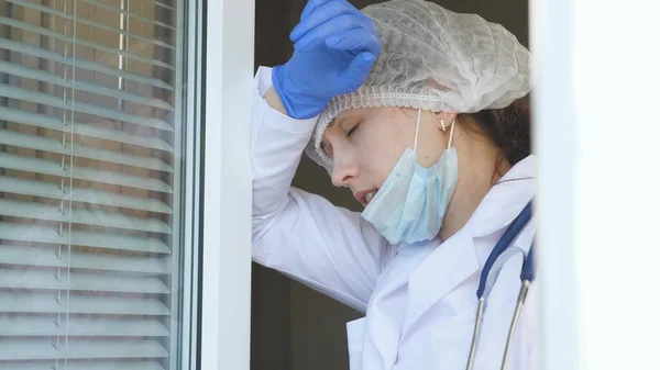 防護服を着た疲れた女性医師が窓に近づき、彼女の医療マスクと帽子を脱ぎ、疲労から目を閉じ、新鮮な空気を呼吸します。病院での医師の勤務日. — ストック写真