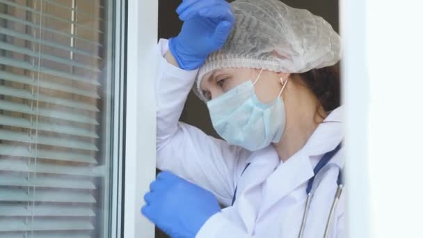 Una doctora cansada en traje protector se acerca a la ventana, se quita la máscara médica y el sombrero, cierra los ojos por fatiga, respira aire fresco. Día de trabajo duro del médico en el hospital . — Vídeos de Stock