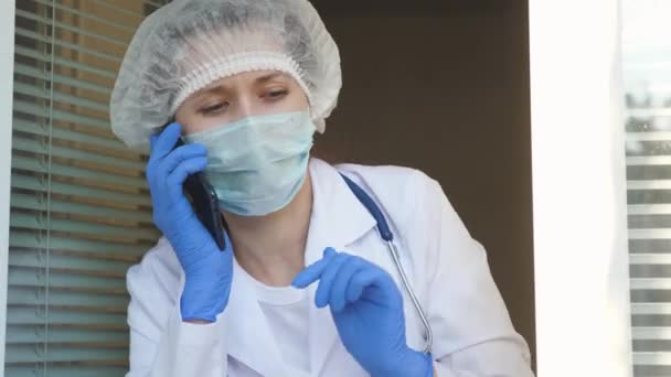 Female doctor in a protective suit looks out window, talking on phone. nurse is waving at window with his hand and talking on smartphone. Doctors working day at hospital. Pandemic coronavirus — Stock Video