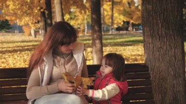 Anne ve bebek sonbahar akçaağaç yapraklarıyla oynuyorlar. Küçük kız ve anne sonbahar parkında bankta oynuyorlar. Mutlu çocukluk kavramı. Çocuklu mutlu bir aile sarı yapraklarla parkta yürüyor.