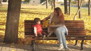 Küçük kız ve anne sonbahar parkında bankta oynuyorlar. Mutlu çocukluk kavramı. Anne ve bebek sonbahar akçaağaç yapraklarıyla oynuyorlar. Çocuklu mutlu bir aile sarı yapraklarla parkta yürüyor.