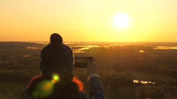 Young female traveler takes photos and videos on her smartphone at dawn, from mountain in rays of beautiful sun. free girl tourist enjoys beautiful views of nature from a high hill. concept of travel — Stock Video