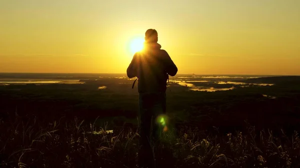 Un viaggiatore con uno zaino si trova ai margini della montagna nei raggi dell'alba, alza le mani in aria e gode della vittoria, del bel sole e del paesaggio. gratis maschio turista viaggia da solo nel parco — Foto Stock