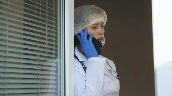 A nurse breathes fresh air by window and talks on smartphone. female doctor in a protective suit looks out the window, talking on phone. Doctors working day at hospital. Pandemic coronavirus