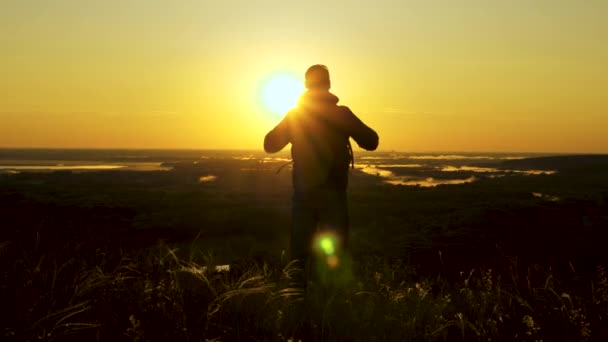 A traveler with a backpack stands on edge of mountain in rays of dawn, raises his hands in air and enjoys the victory, beautiful sun and landscape. free male tourist travels alone in park — Stock Video
