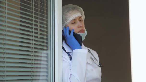 Una enfermera respira aire fresco por la ventana y habla en el teléfono inteligente. doctora con traje protector mira por la ventana, hablando por teléfono. Doctores trabajando en el hospital. Coronavirus pandémico — Vídeo de stock