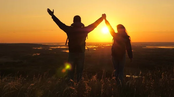 Viajeros, un hombre y una mujer con mochila van al borde de la montaña en rayos del amanecer, levantan las manos en el aire y disfrutan de la victoria, el hermoso sol y el paisaje. Los turistas libres viajan. trabajo en equipo de negocios —  Fotos de Stock