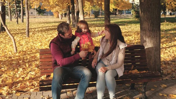 Papá, hija pequeña y mamá juegan en el parque de otoño en el banco. concepto de la infancia feliz. bebé, madre y padre están jugando con hojas de arce de otoño. Familia feliz con un niño camina en un parque de la ciudad . — Foto de Stock