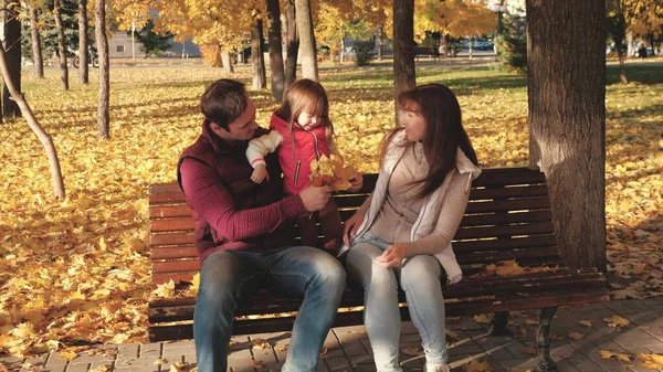 Pappa, lilla dotter och mamma spelar i höstparken på bänk. begreppet lycklig barndom. Baby, mor och far leker med höstlöv. Lycklig familj med ett barn promenader i en stadspark. — Stockfoto