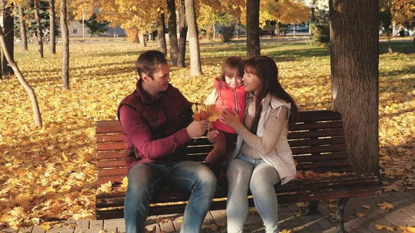 Pappa, lilla dotter och mamma spelar i höstparken på bänk. begreppet lycklig barndom. Baby, mor och far leker med höstlöv. Lycklig familj med ett barn promenader i en stadspark. — Stockfoto