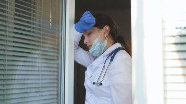 Koruyucu kıyafetli yorgun kadın doktor pencereye doğru yürür, tıbbi maskesini çıkarır, gözlerini yorgunluktan kapatır, temiz hava soluyarak nefes alır. Doktor bugün hastanede çok çalışıyor. salgın covid 19.