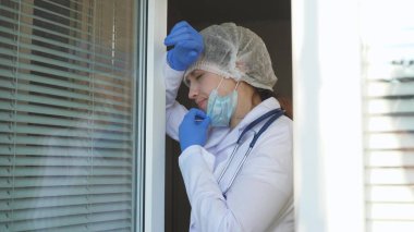 Koruyucu kıyafetli yorgun kadın doktor pencereye doğru yürür, tıbbi maskesini çıkarır, gözlerini yorgunluktan kapatır, temiz hava soluyarak nefes alır. Doktor bugün hastanede çok çalışıyor. salgın covid 19.