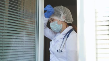 Koruyucu kıyafetli yorgun kadın doktor pencereye doğru yürür, tıbbi maskesini çıkarır, gözlerini yorgunluktan kapatır, temiz hava soluyarak nefes alır. Doktor bugün hastanede çok çalışıyor. salgın covid 19.