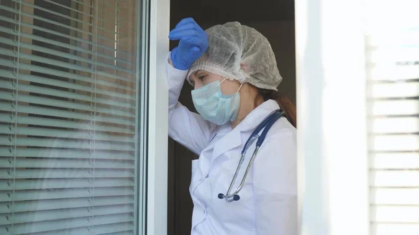 防護服を着た疲れた女性医師が窓まで歩き彼女の医療マスクを脱ぎ疲れから目を閉じて新鮮な空気を呼吸する。医者は病院で一生懸命働く。伝染病のコヴィド19. — ストック写真